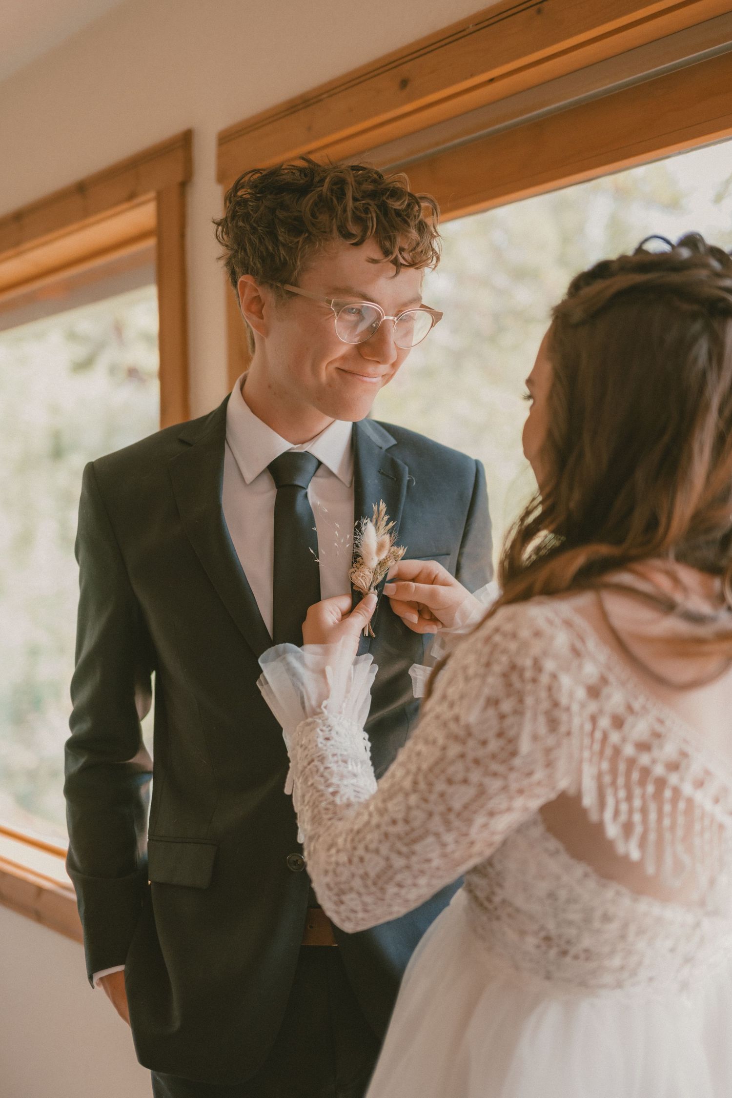 A bride adjusts the boutonniere on her groom, who looks at her with a smile.