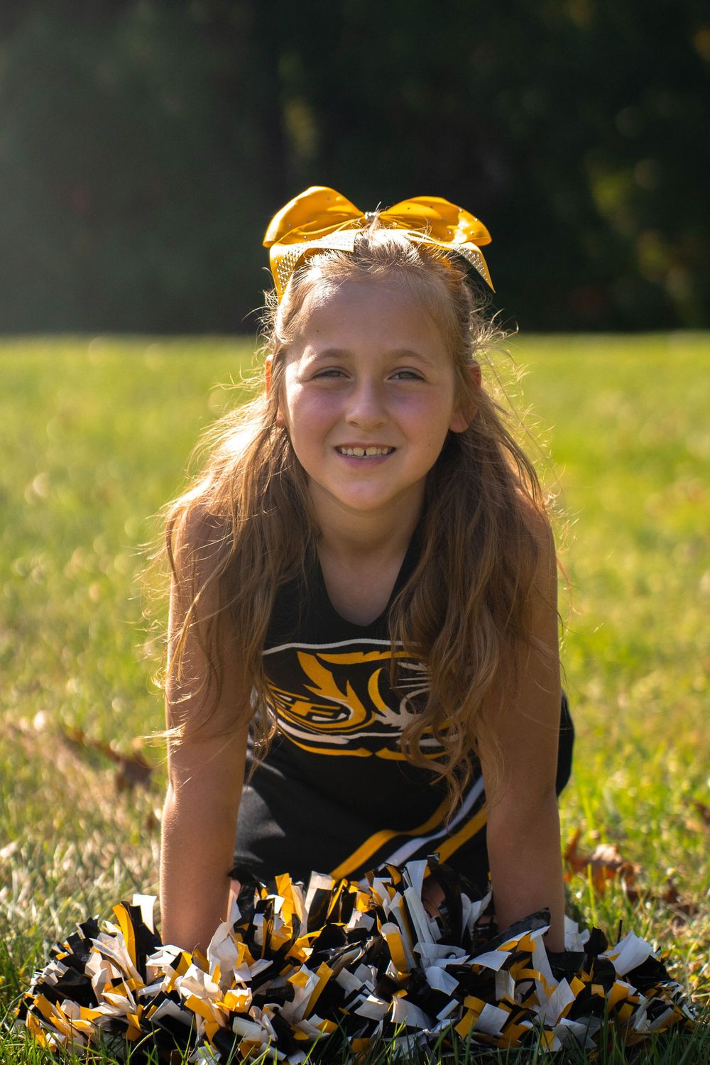 Smiling young cheerleader in black and gold uniform sitting on grass in sunlight.