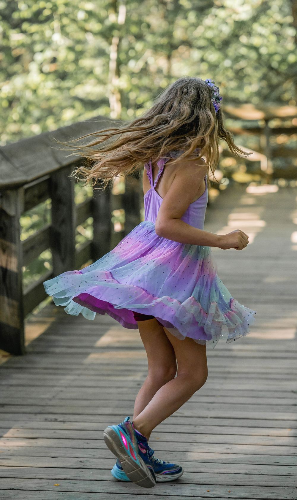 A young girl in a purple tulle fairy dress twirls on a wooden bridge in a forest setting.