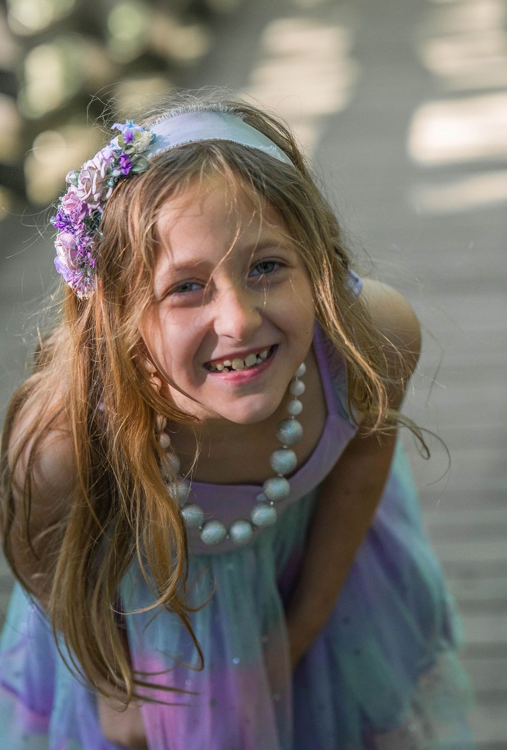 A smiling young girl wearing a purple fairy dress and floral crown accessory with a pearl necklace.
