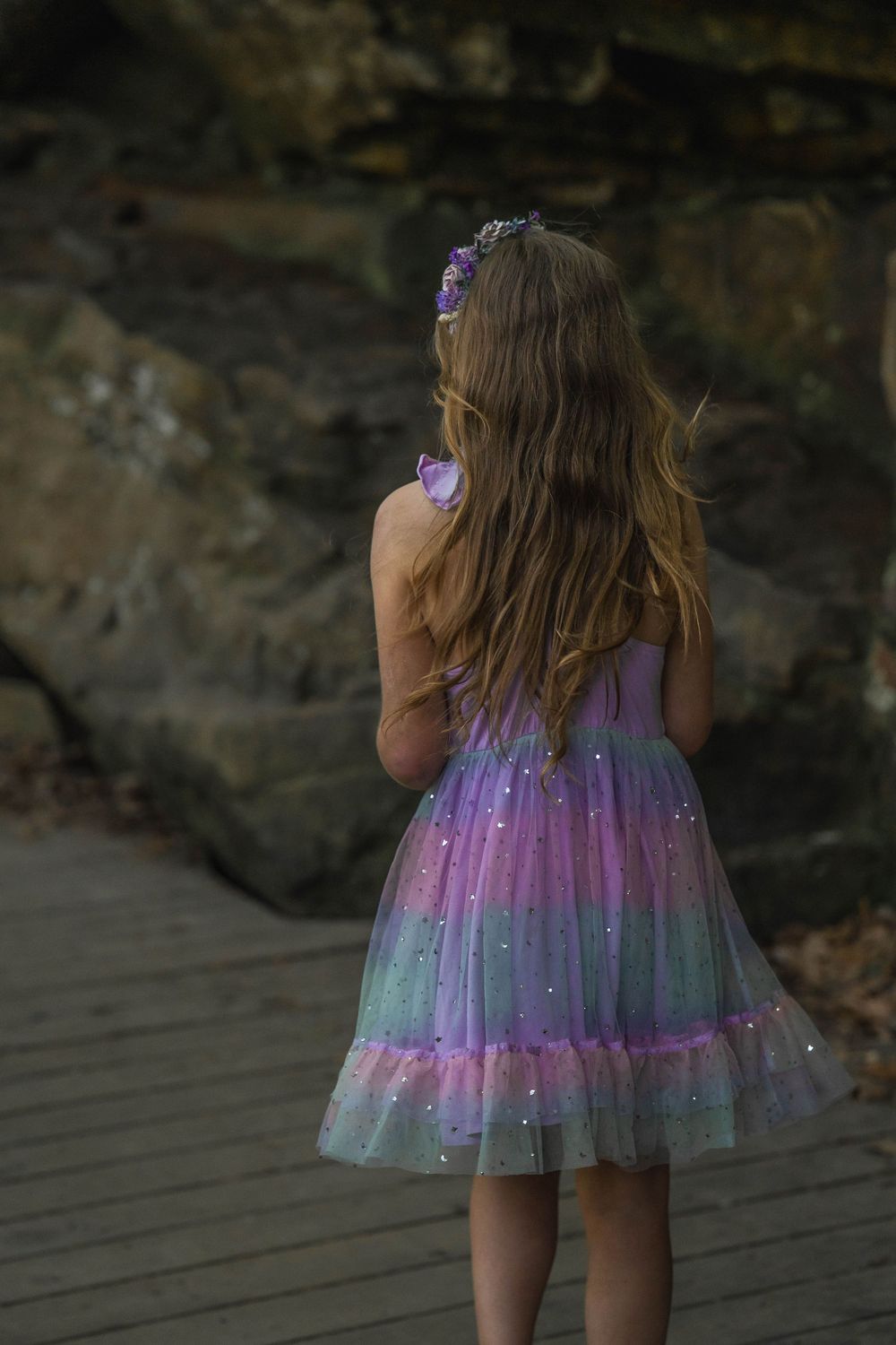 A young girl in a sparkling purple rainbow tulle dress stands on a wooden path facing rocky cliffs.