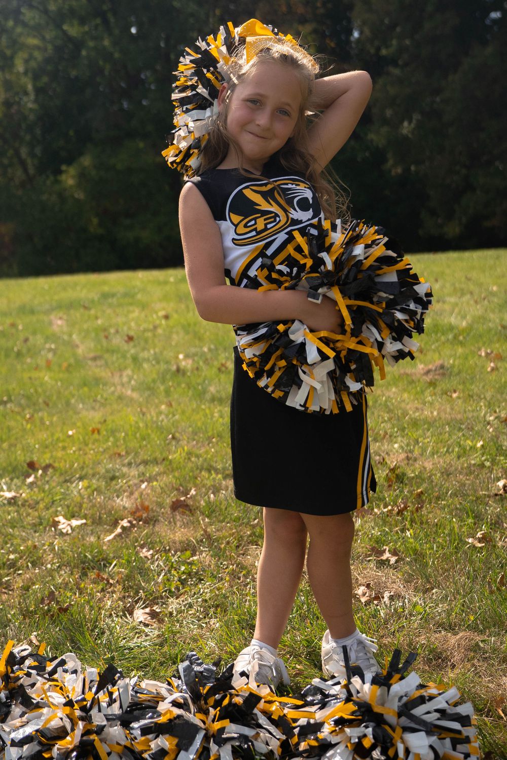 Smiling young cheerleader in black and gold uniform poses with pom-poms on a sunny outdoor field.