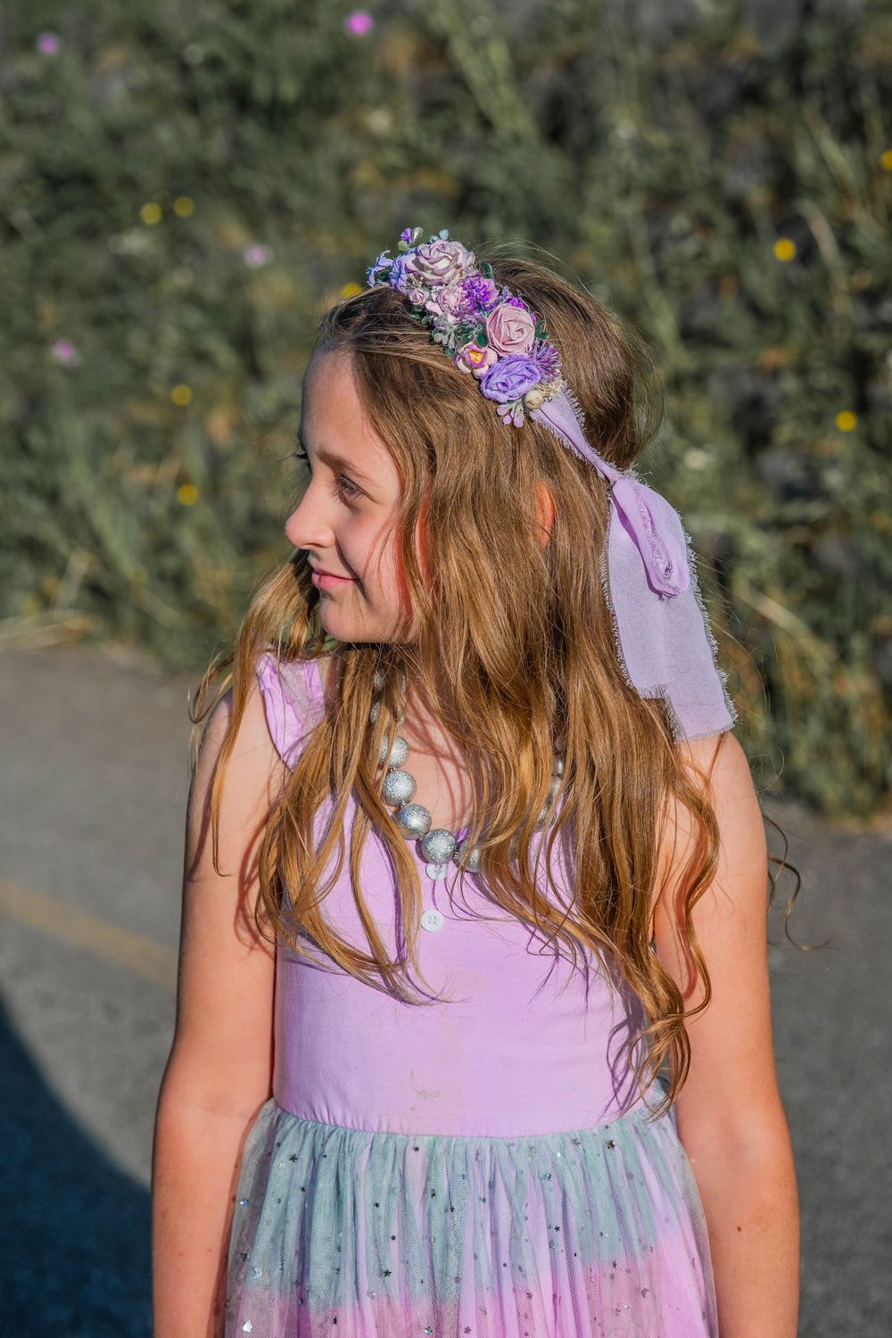 A young girl wearing a lavender fairy dress and floral crown headpiece looks sideways outdoors.