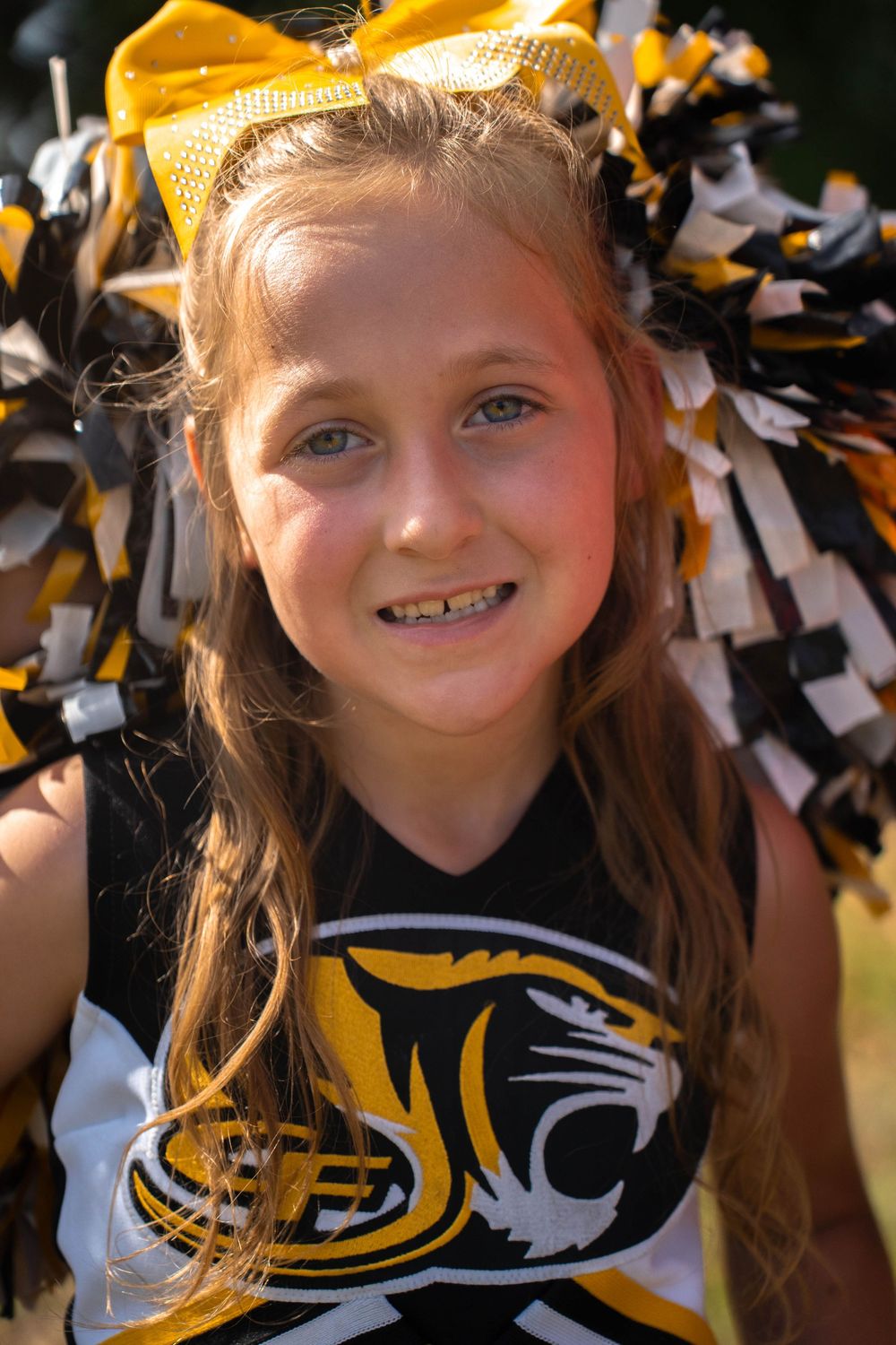 Close-up portrait of a young cheerleader smiling in black and gold uniform with yellow bow and pom-poms.