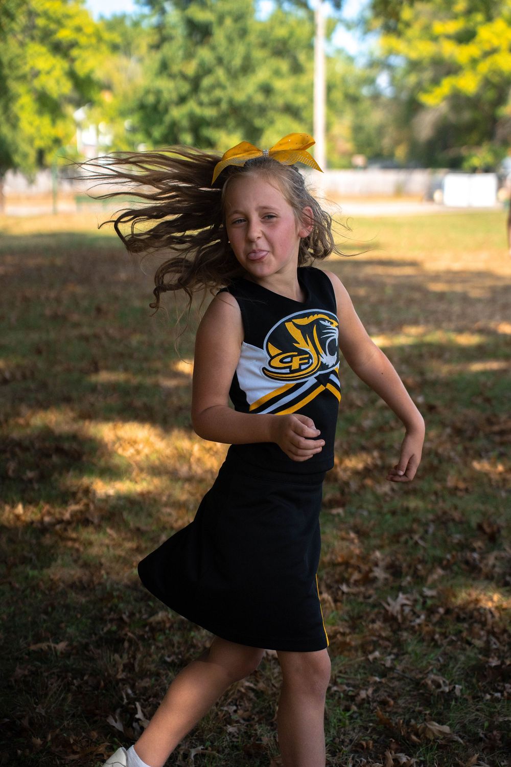 Young girl in black cheerleader uniform spins with hair flying outdoors in a park setting.