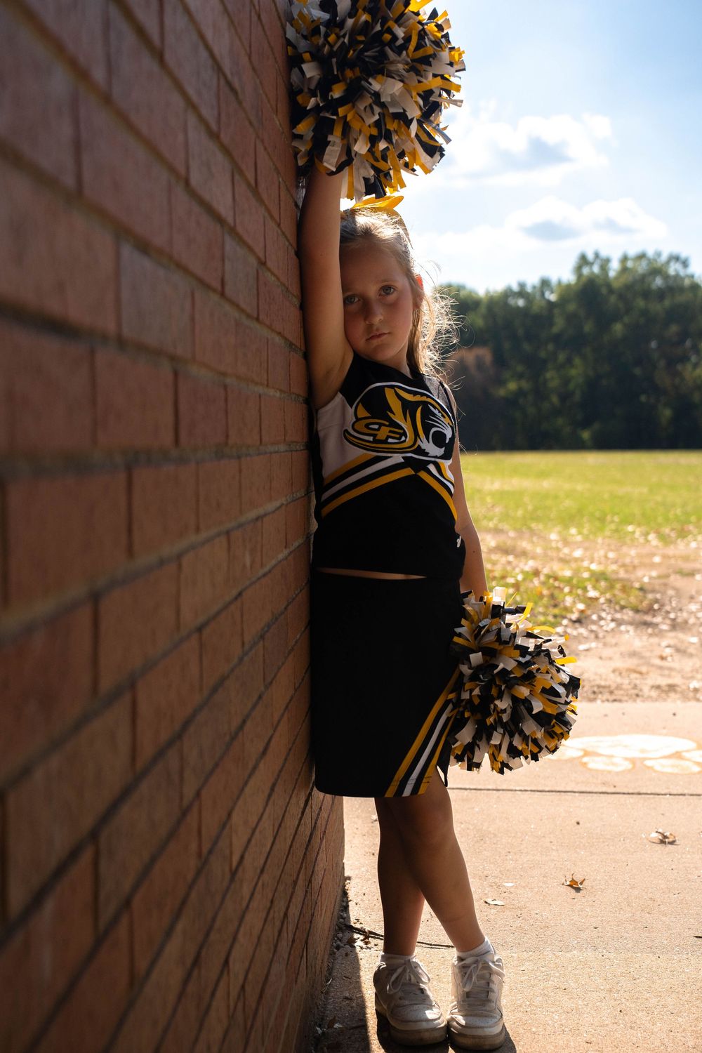 Young cheerleader in black and gold uniform leans against brick wall holding pom-poms outdoors.