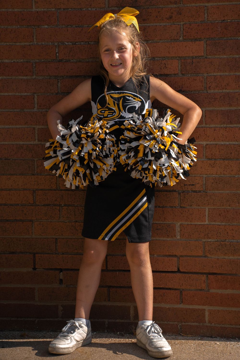 Smiling young cheerleader in black and gold uniform stands against brick wall holding colorful pom-poms.