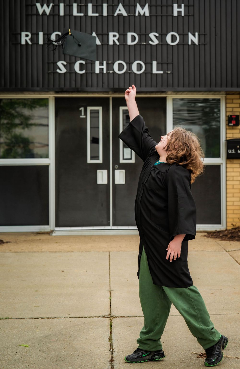 Graduate in black cap and gown tossing mortarboard outside William H. Richardson School.