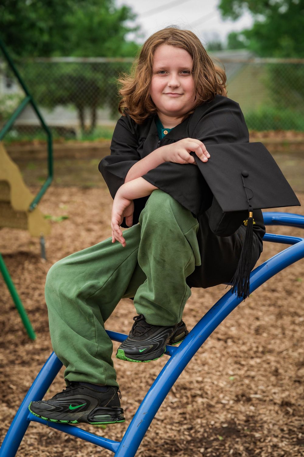 Young boy with long hair sitting on playground equipment holding graduation cap and gown.