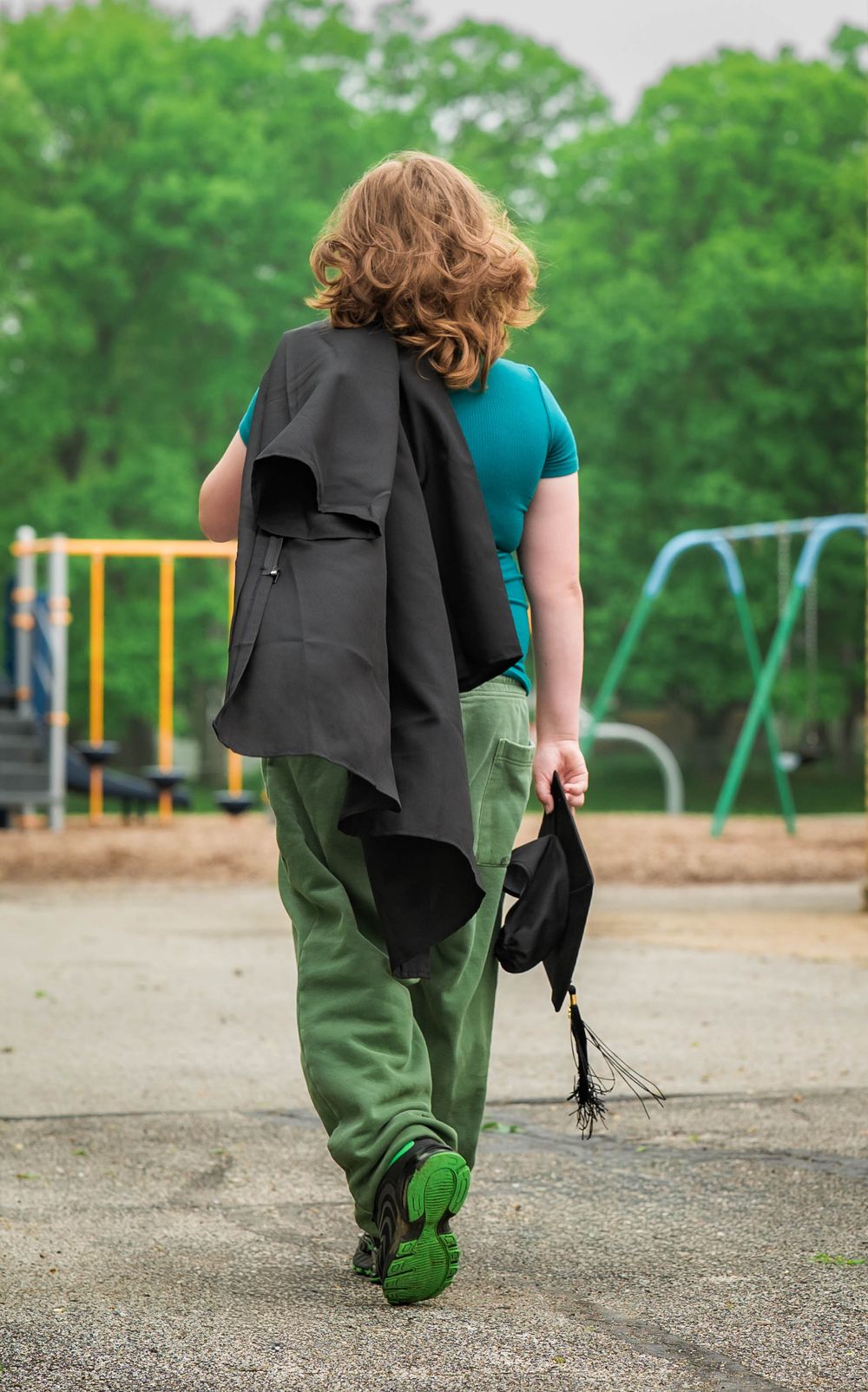 Boy walking through playground carrying black graduation gown and cap from behind.