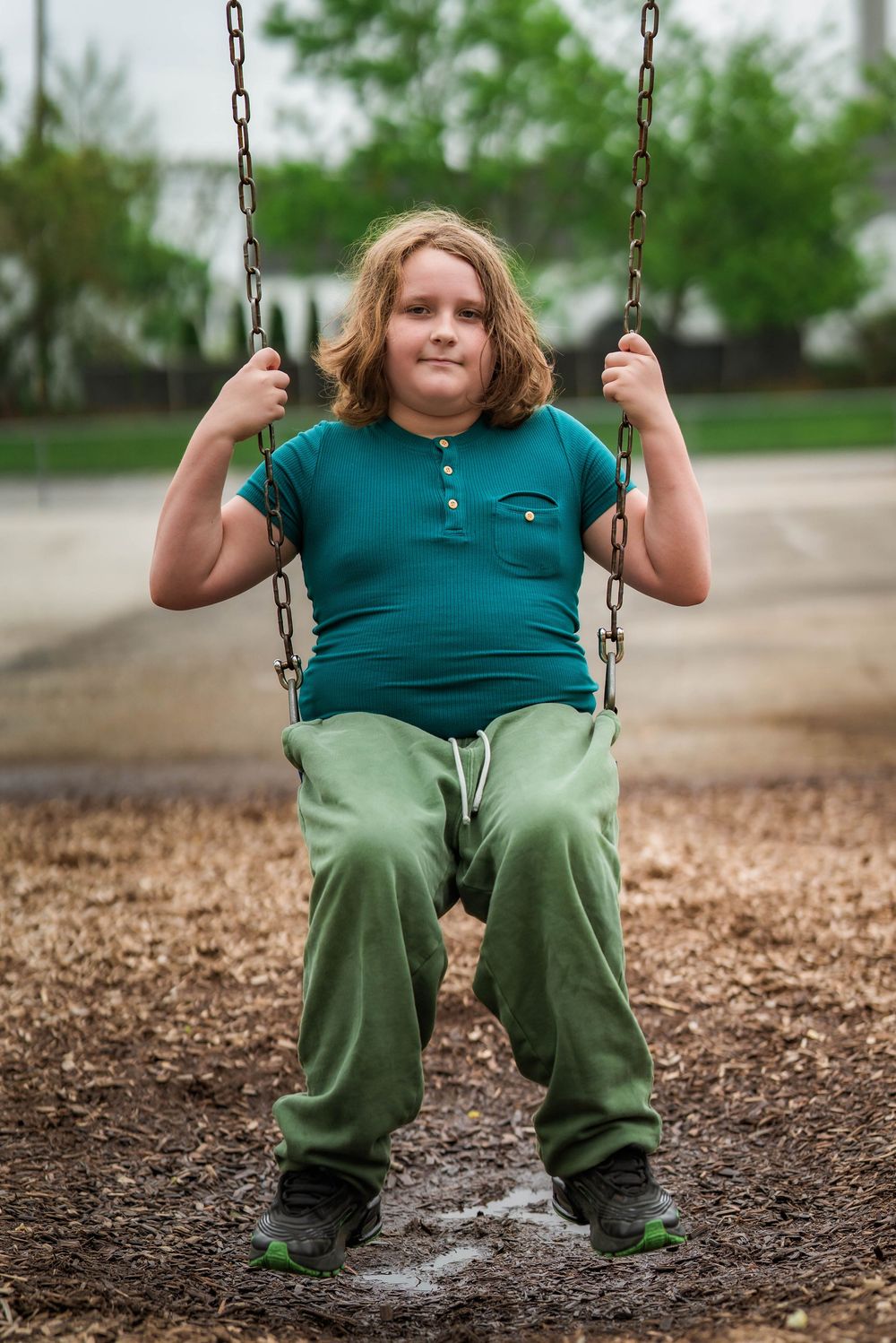 A child with long auburn hair sits on a chain swing at a playground wearing a teal shirt and green pants.