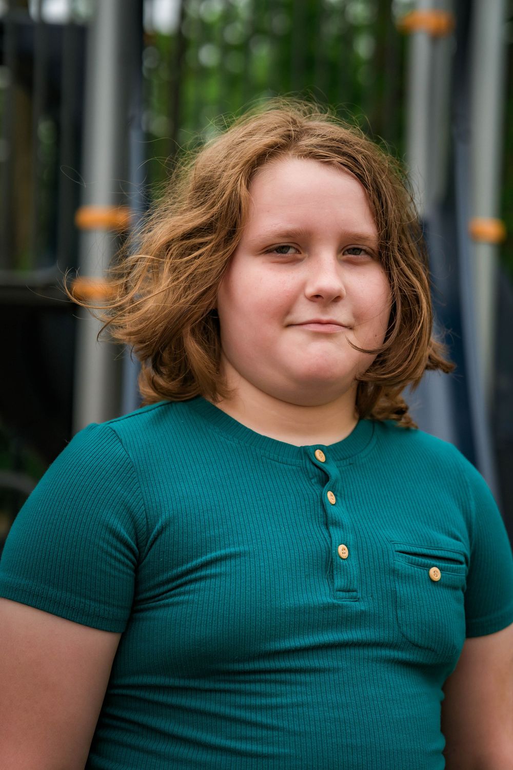 A young child with wavy auburn hair wearing a teal henley shirt smiles softly at a playground.