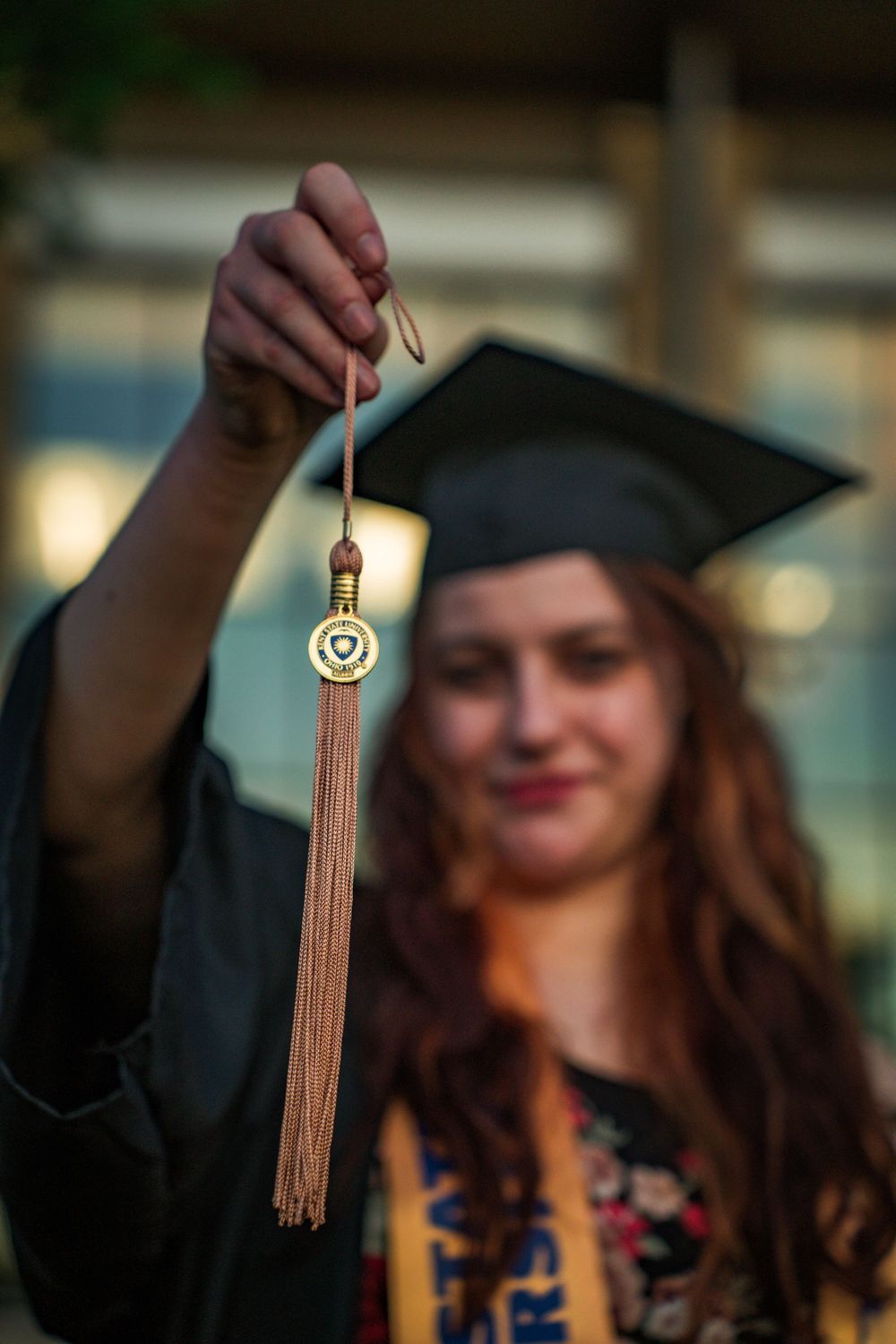Graduate holds up gold tassel medallion in focus with blurred graduate in cap and gown behind.