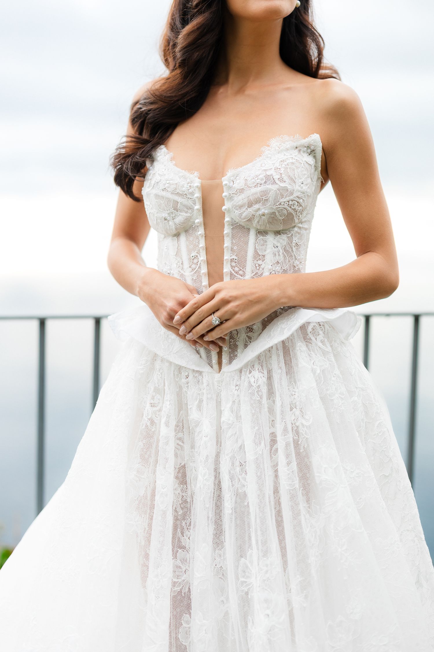 Bride in strapless lace corset wedding gown with deep V neckline holding hands at ocean overlook.