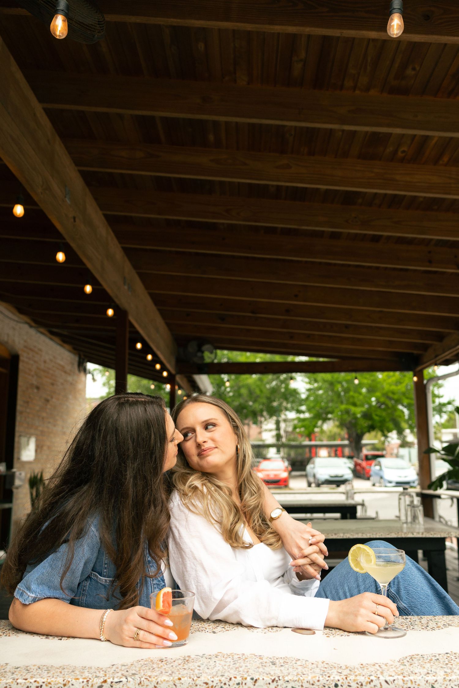 Two women enjoying cocktails at an outdoor bar patio, sharing an intimate moment under warm string lights.