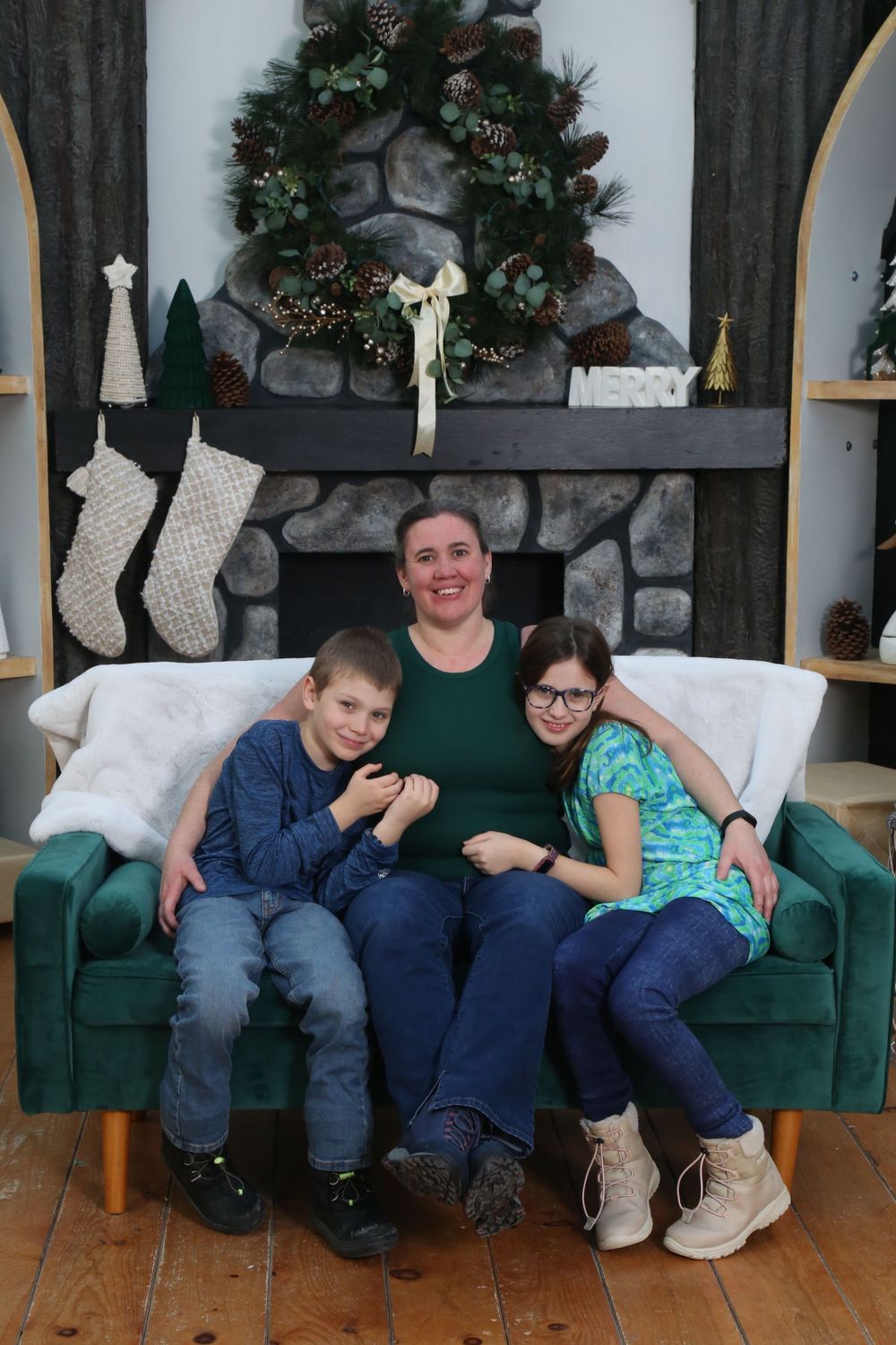 Family of three poses on green sofa before decorated fireplace with Christmas stockings and wreath.