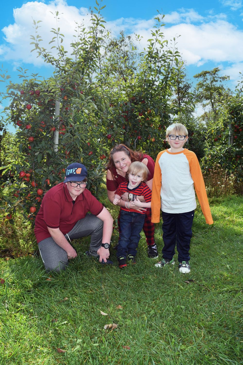 Family of four posing in an apple orchard on a sunny day, with red apple trees and green grass in the background.