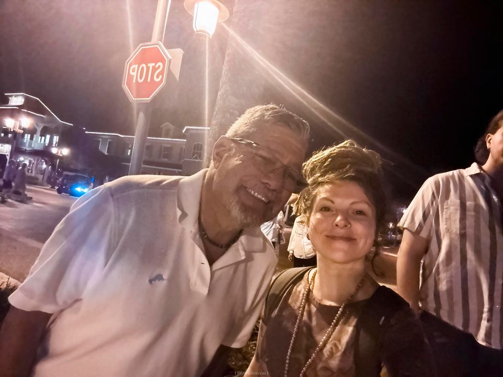 Two people smiling together outdoors at night near a stop sign on a city street.