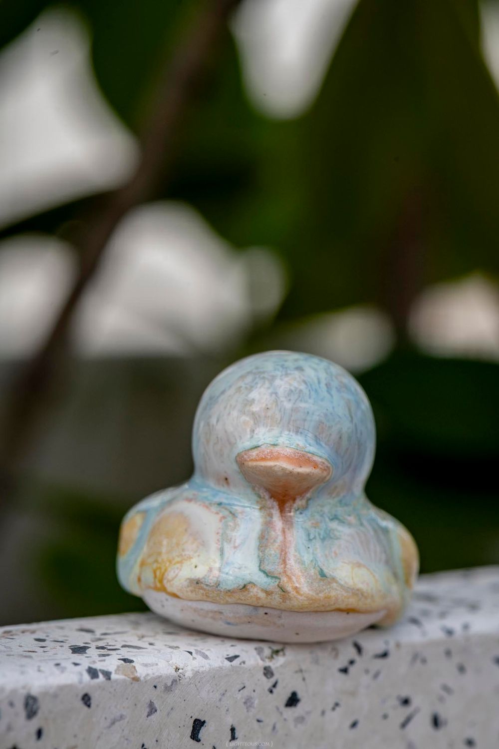 A weathered ceramic duck figurine with blue and peach tones rests on a terrazzo surface outdoors.