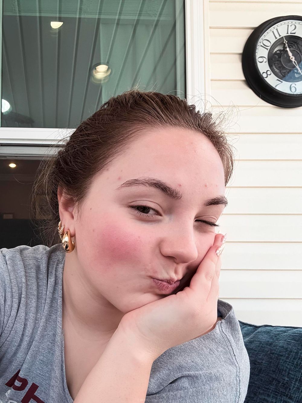 Young woman with rosy cheeks and gold hoop earrings making a pouty face while resting her chin on her hand outdoors.