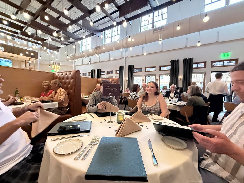 Busy upscale restaurant dining room with guests reviewing menus at white tablecloth tables under high ceilings.