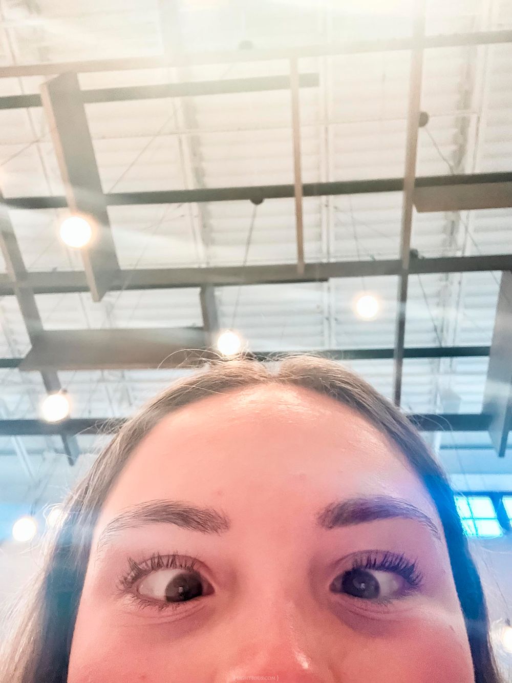 Close-up selfie of a young woman with bold eyebrows and mascara, looking up at an industrial ceiling with lights.