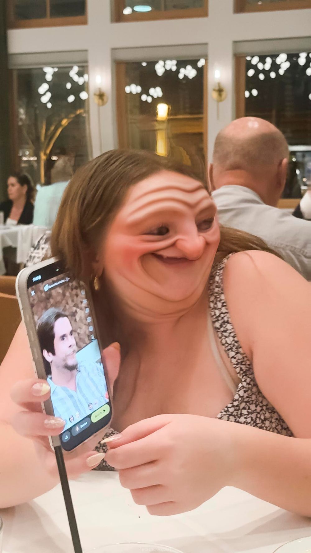 Woman with distorted face filter holds phone showing man's photo while sitting in a restaurant.