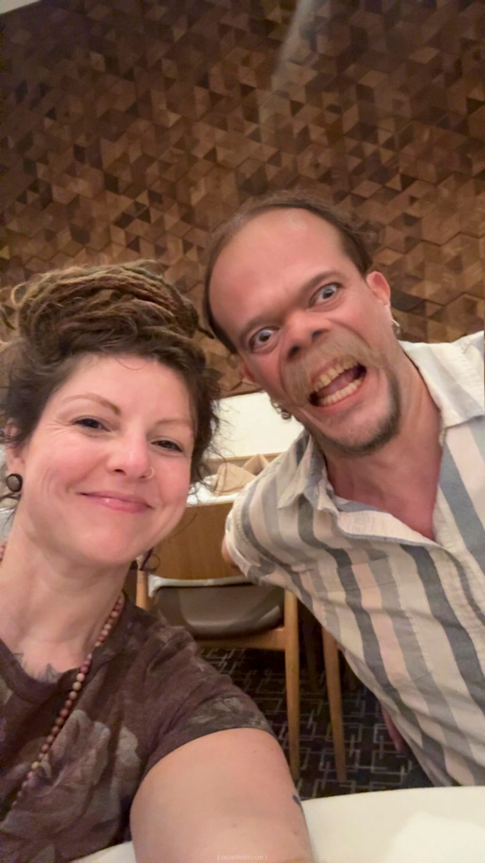 Two smiling adults taking a selfie together in a warmly lit restaurant with a geometric wood panel wall.