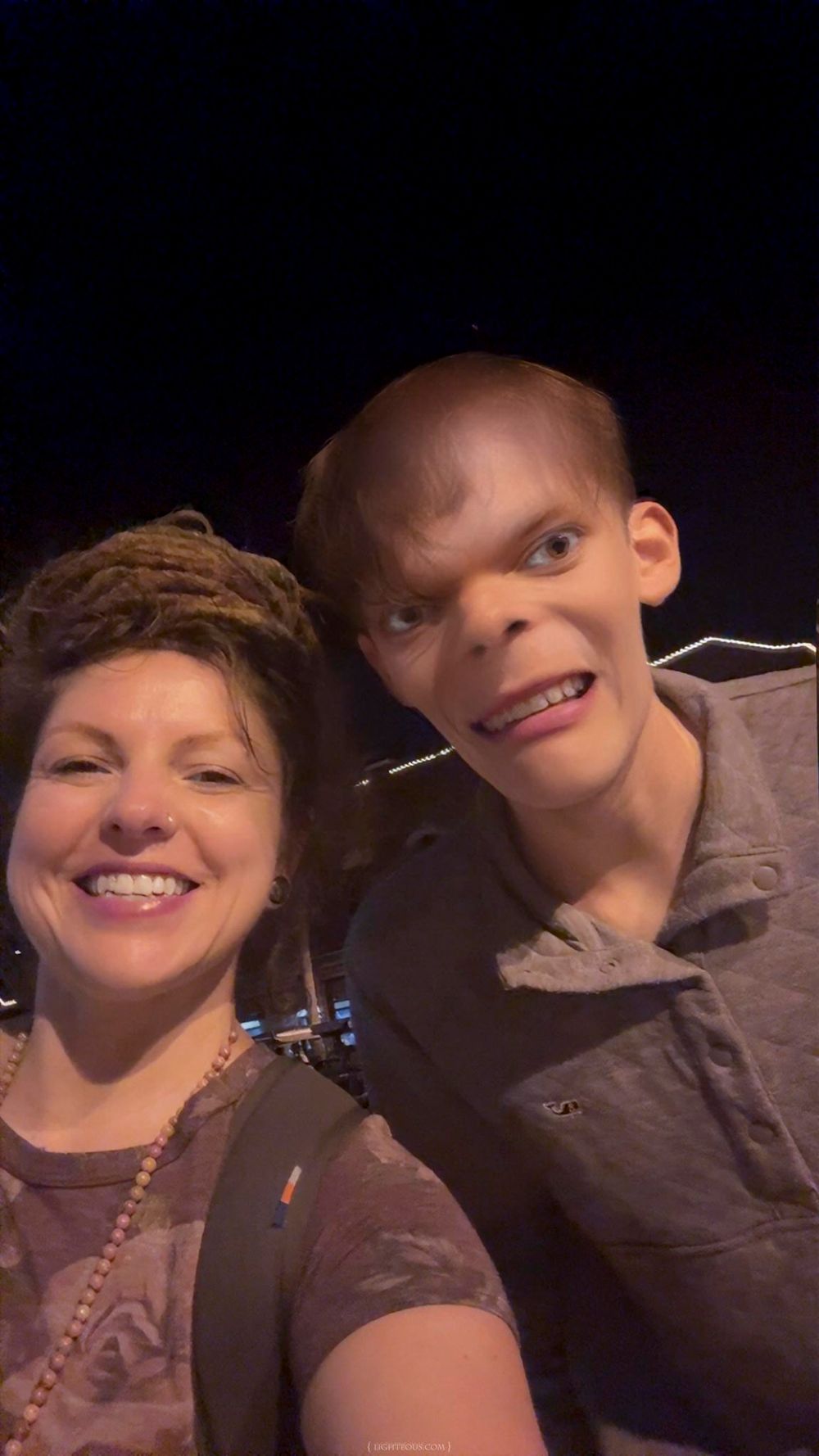 Two people smiling together for a selfie in a dimly lit venue, woman with dreadlocks and man in gray shirt.