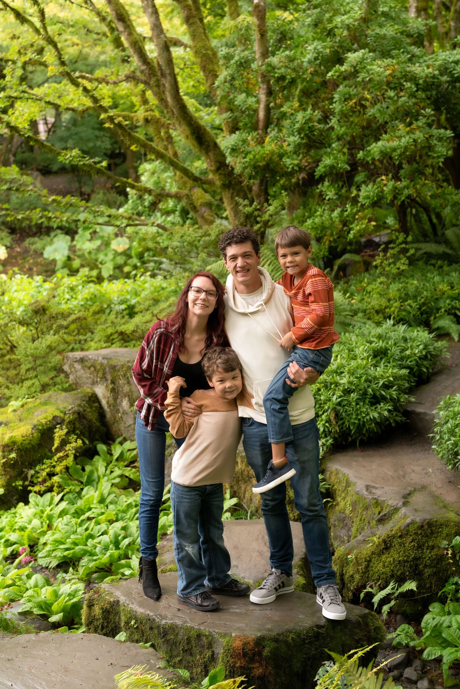 Happy family of four posing on stone steps surrounded by lush green garden foliage.