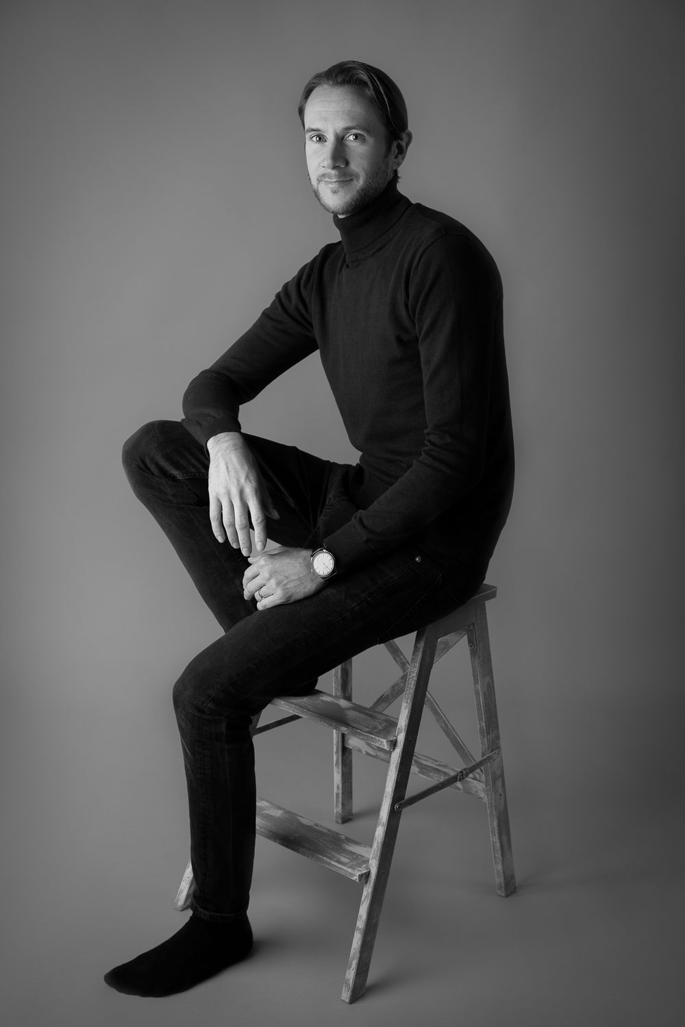 Black and white portrait of a man in turtleneck sitting on wooden ladder stool, looking at camera.