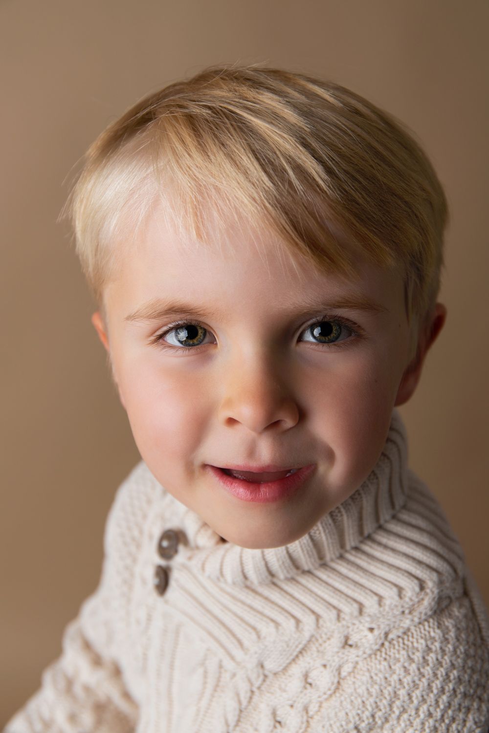 Smiling blonde toddler boy in cream cable-knit sweater poses for portrait against beige background.