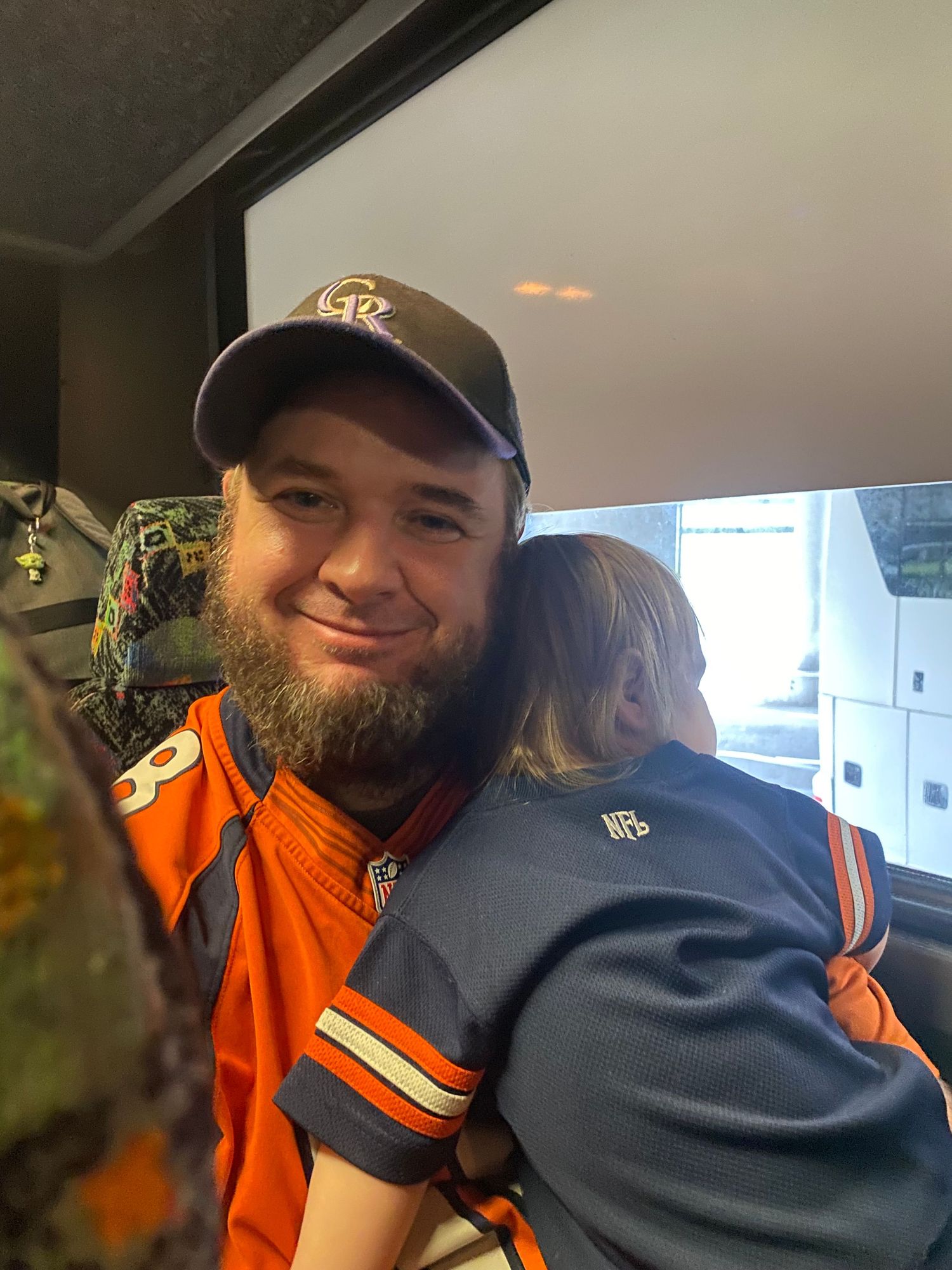 Man in Denver Broncos jersey and Colorado Rockies cap hugs a child wearing a Bears jersey inside a vehicle.