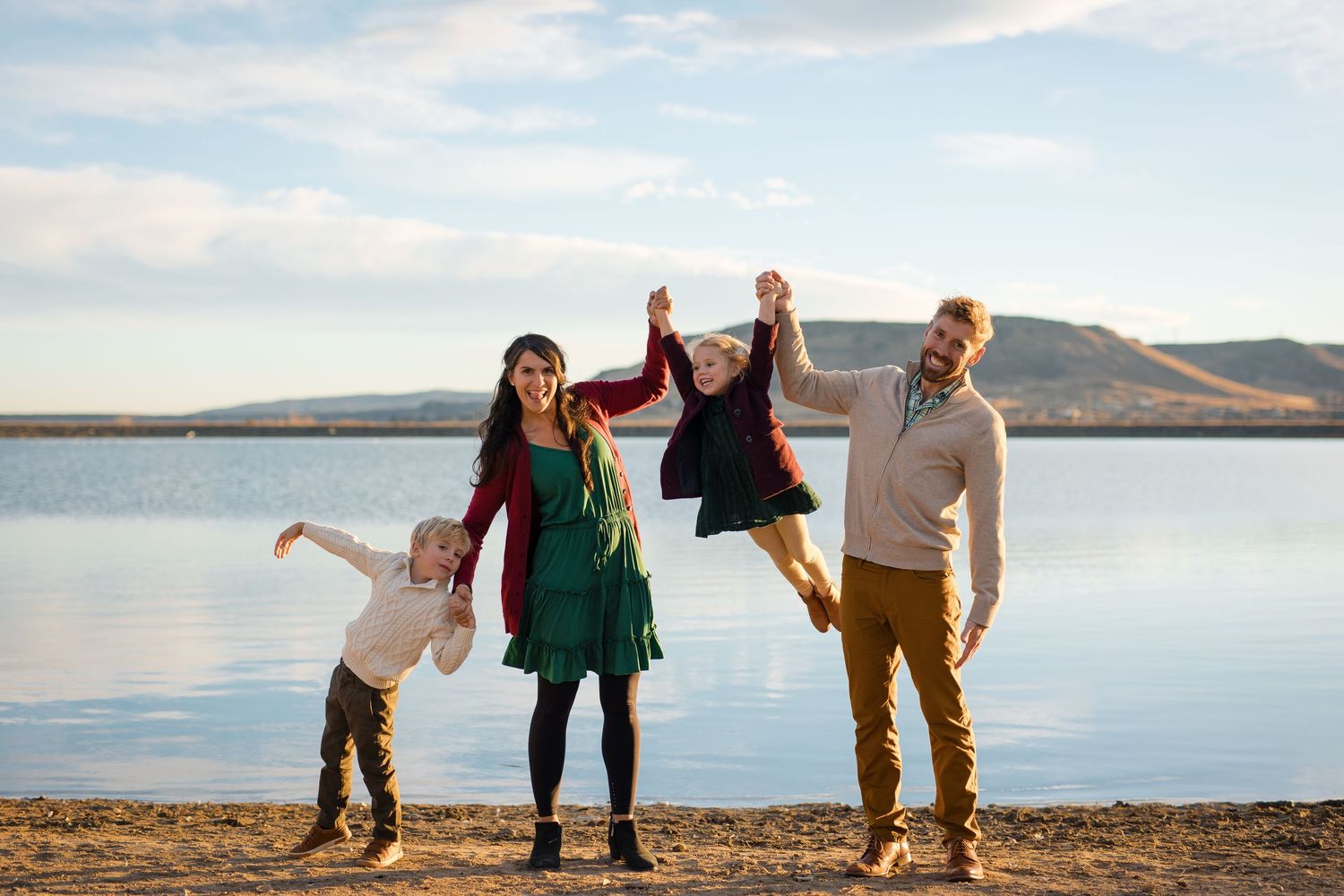 A family enjoys a playful moment together on the shoreline of a serene lake with mountains in the background.