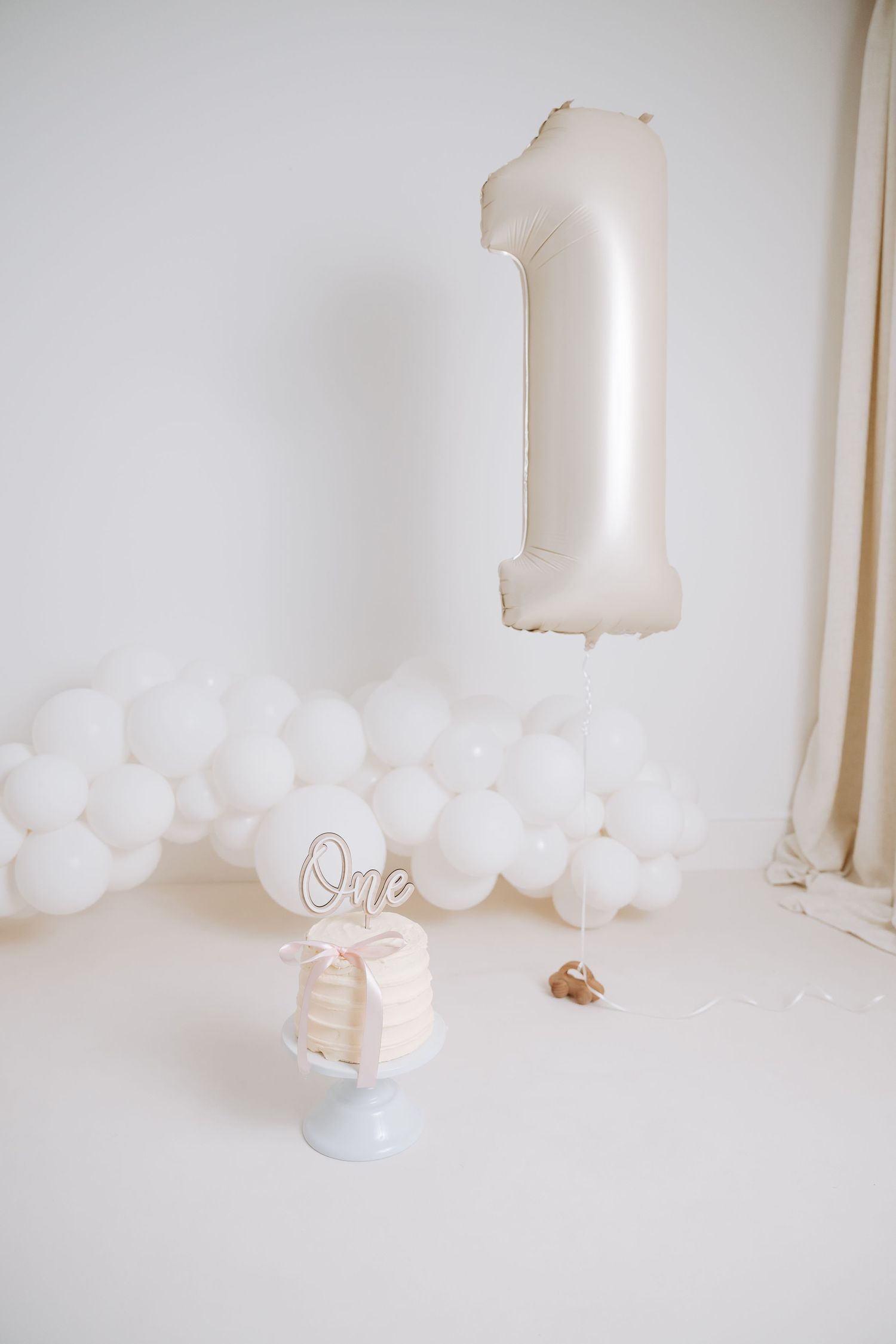 Minimalist white party decor setup with balloons and a cake stand against a light neutral background.