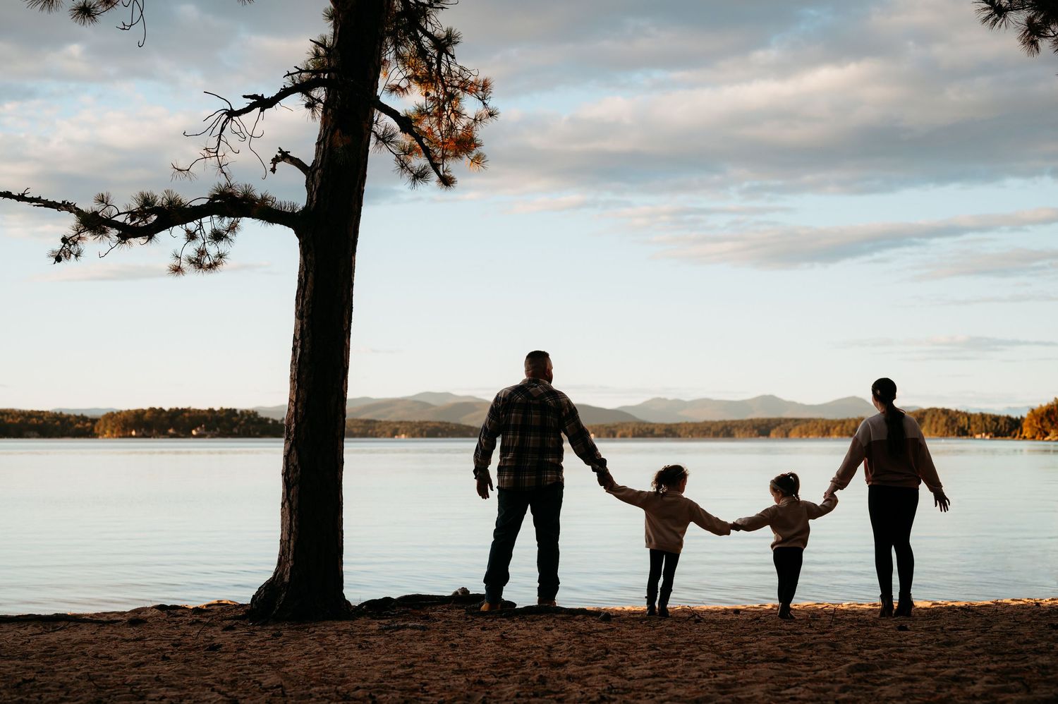 a dad and mom hold hands with their small girls. They are in shadow. The lake and mountains is behind them.