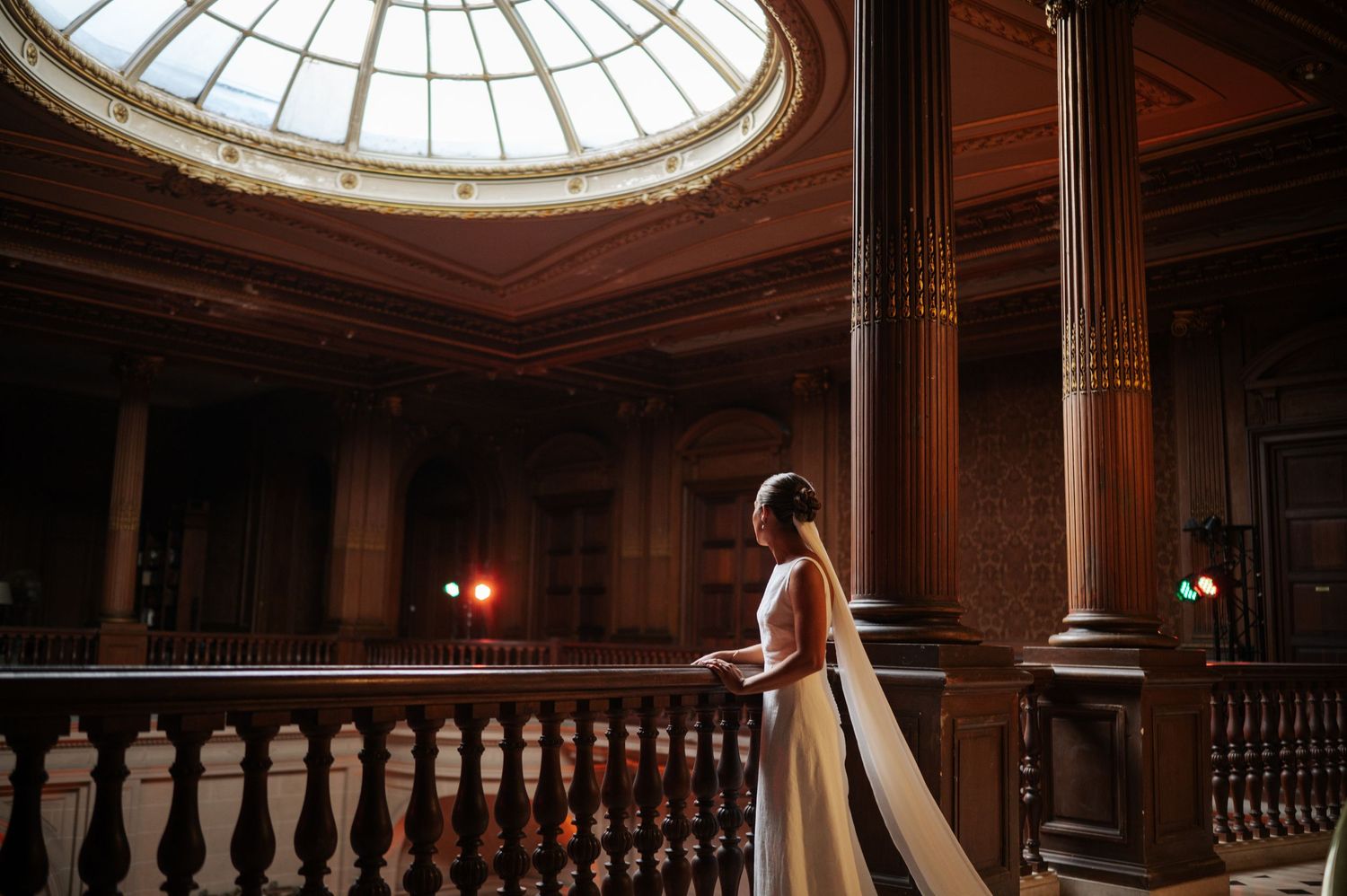 Bride in a white gown standing on a balcony inside an elegant domed historic building.