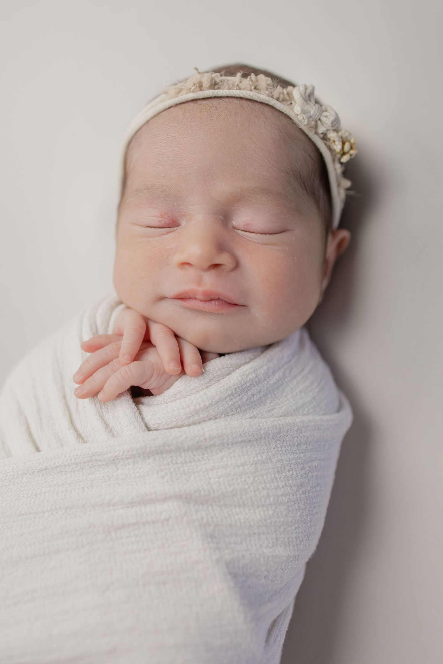 Peaceful newborn baby girl swaddled in white with floral headband sleeping.