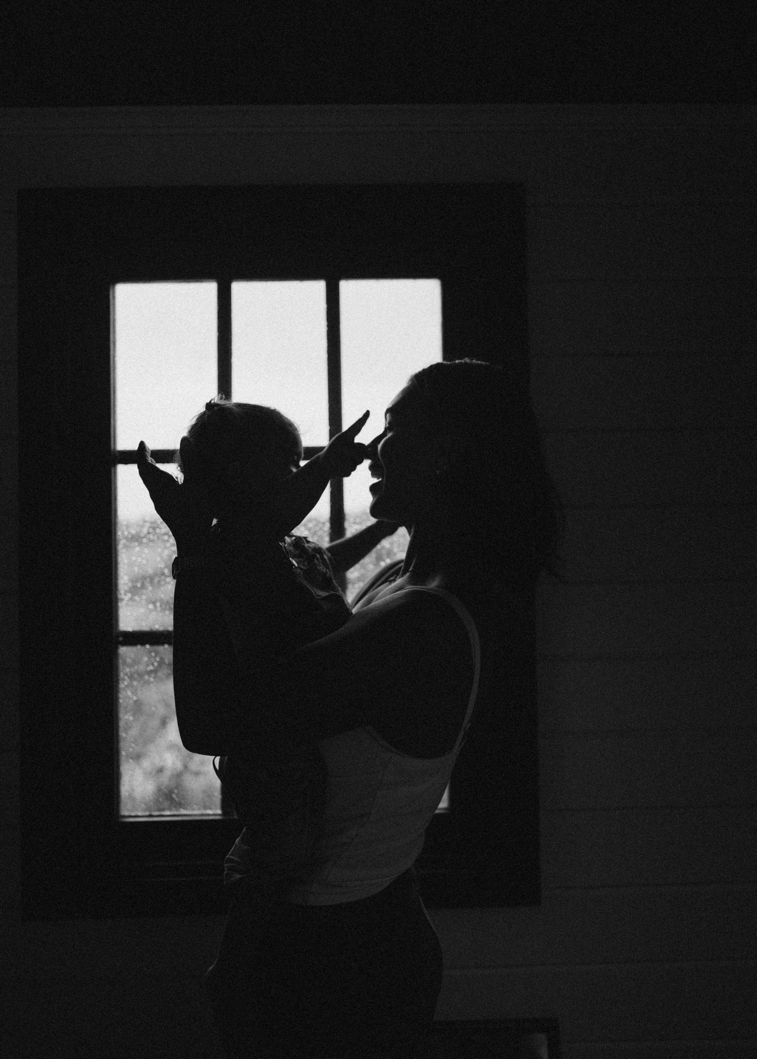 Silhouette of mother lifting baby near window in black and white photo in New Zealand.