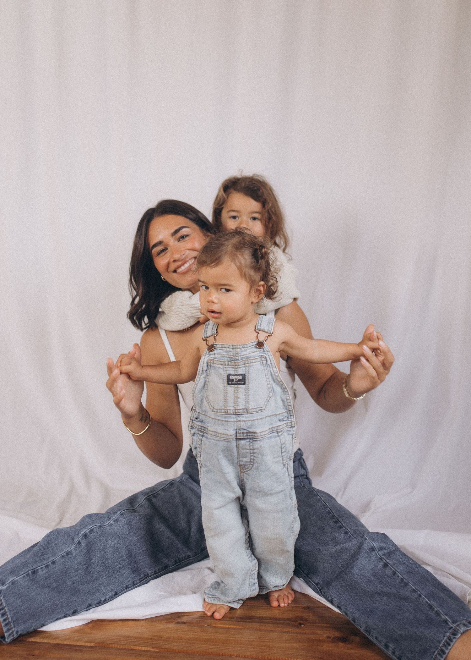 Mother sitting on floor holding toddler in denim overalls while older child stands behind against white backdrop in New Zealand.