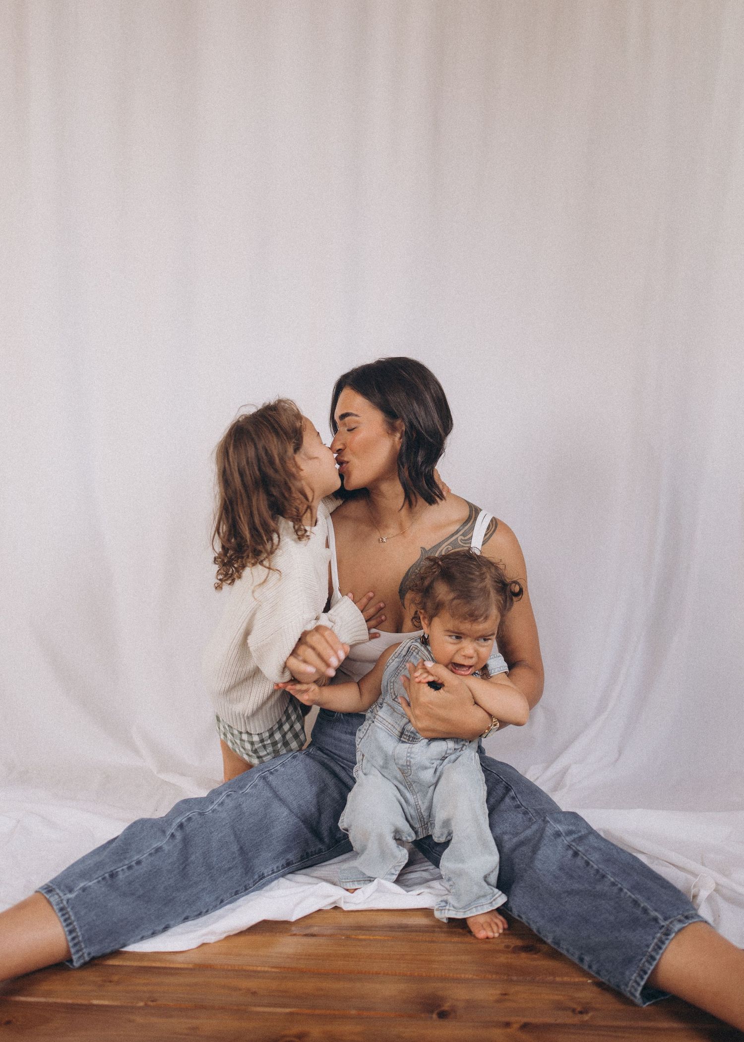 Mother sitting on floor kissing young child while holding baby in denim overalls against white backdrop in New Zealand.