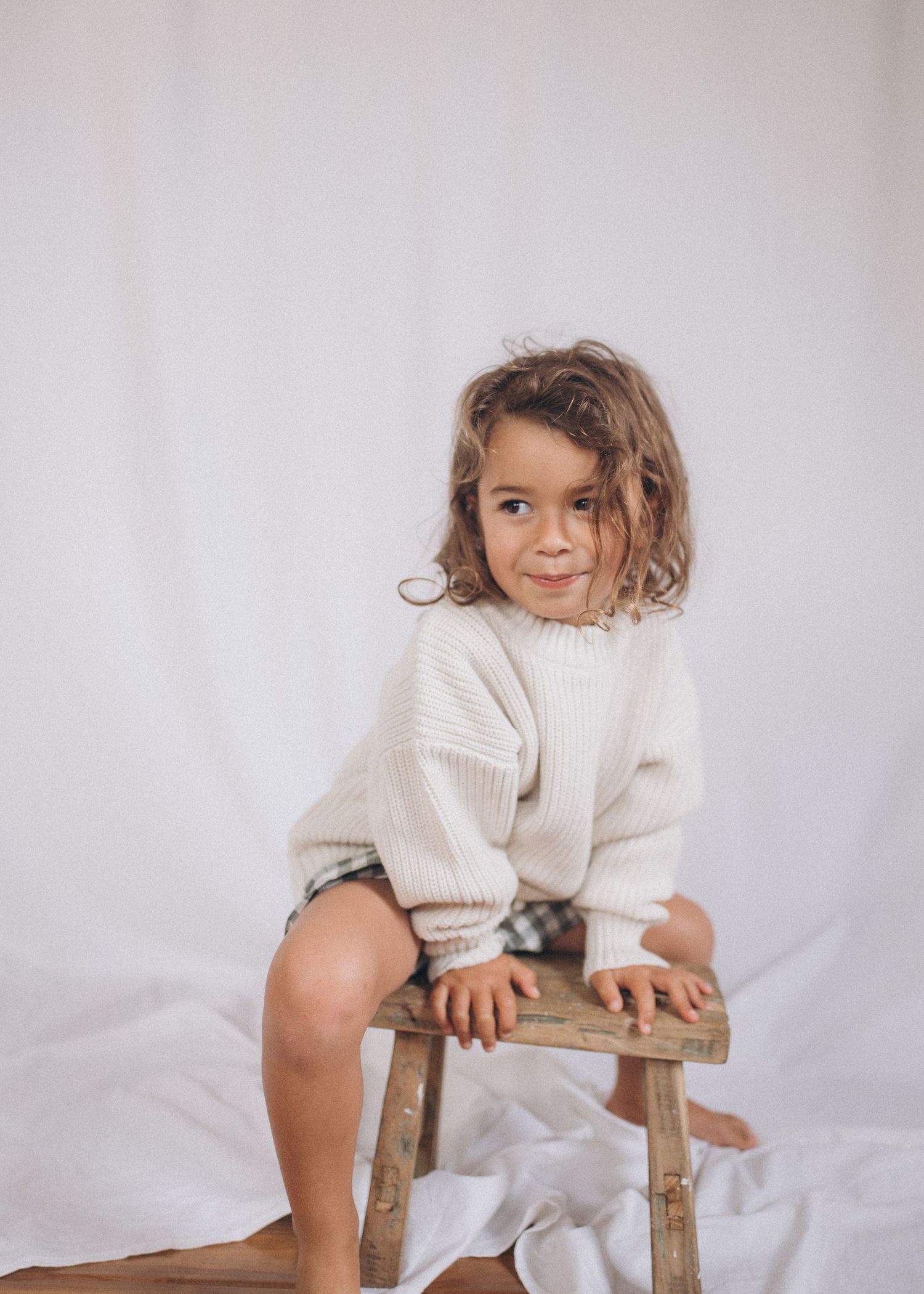 Curly-haired toddler in cream knit sweater sitting on rustic wooden stool in New Zealand.