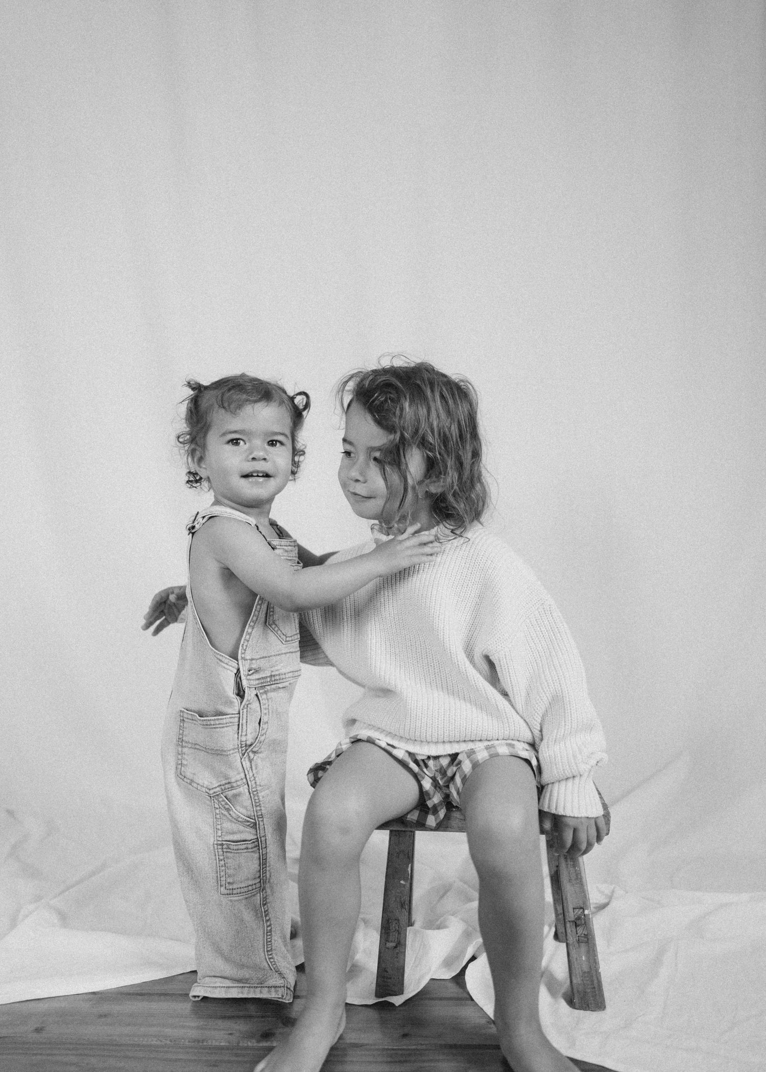Two young sisters posing together in black and white photo, younger in overalls, older in knit sweater in New Zealand.