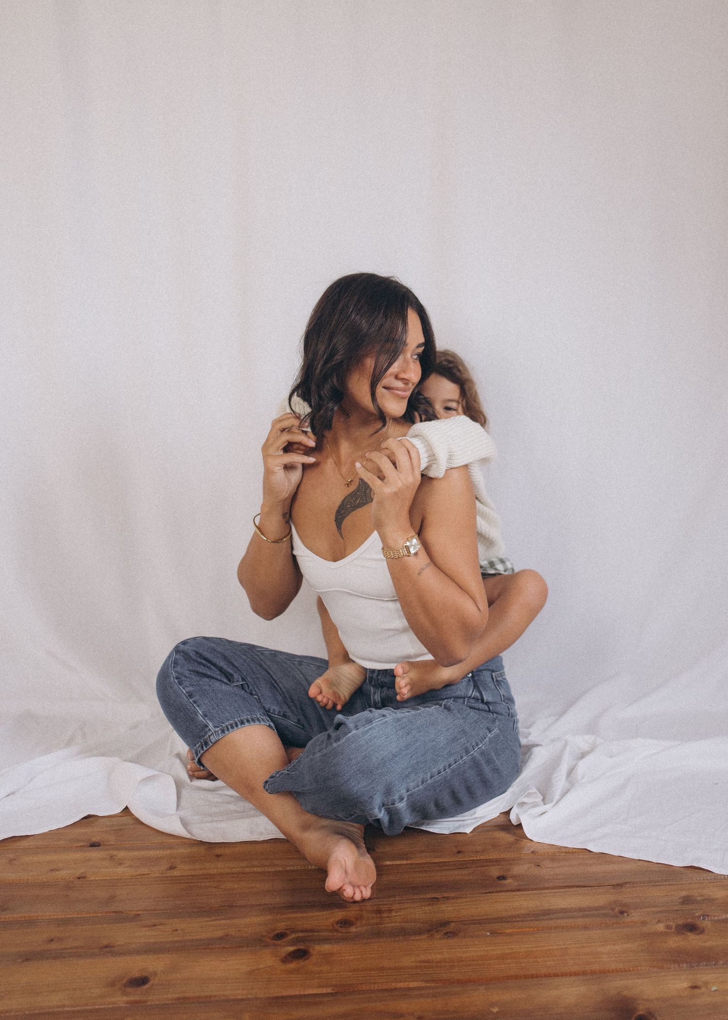 Mother sitting cross-legged on wood floor while child hugs her from behind in New Zealand.