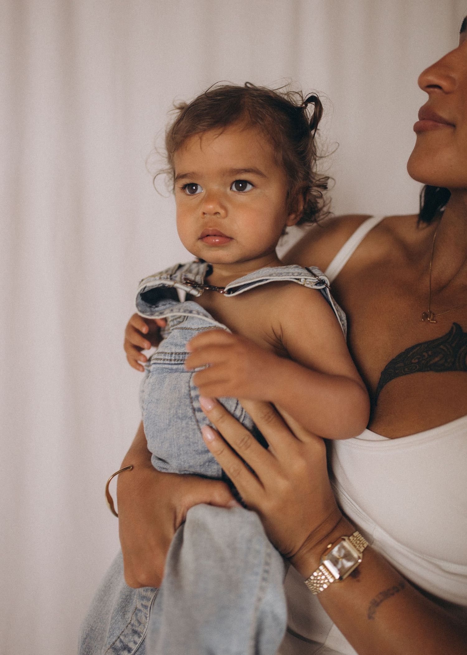 Mother in white top holding baby in denim overalls against white backdrop in New Zealand.