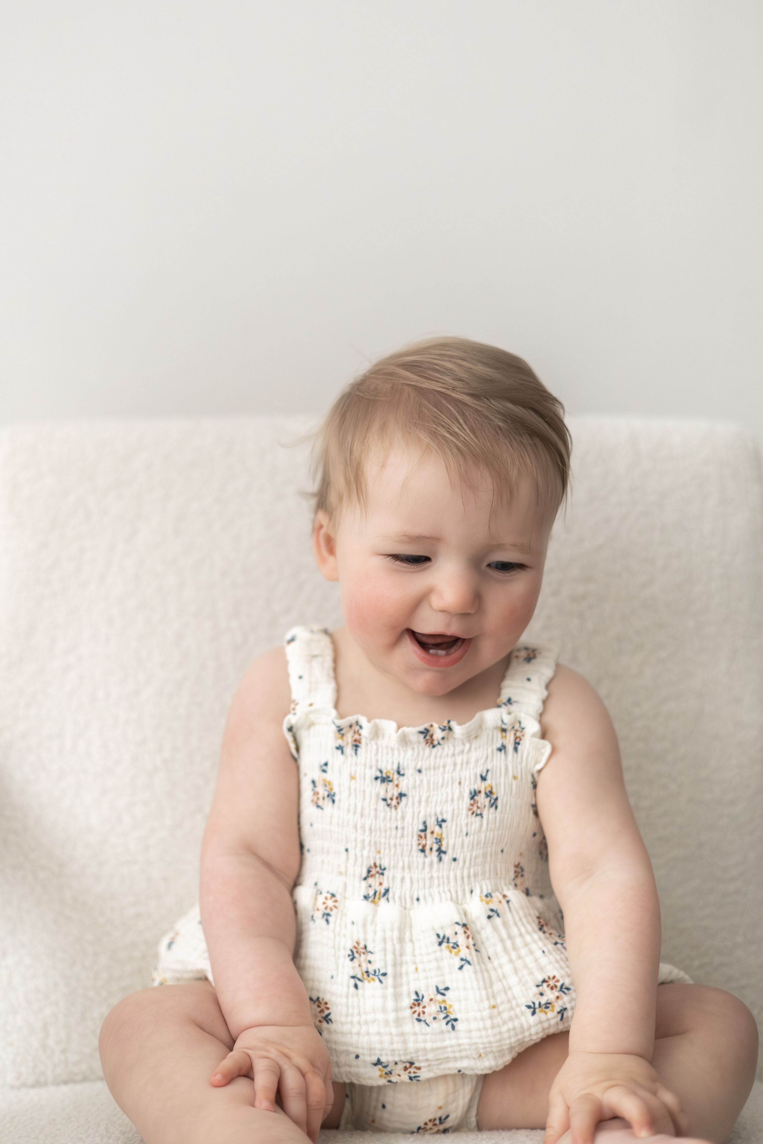 Happy baby girl in floral romper sitting on white chair, laughing with joy first birthday photoshoot Nottingham