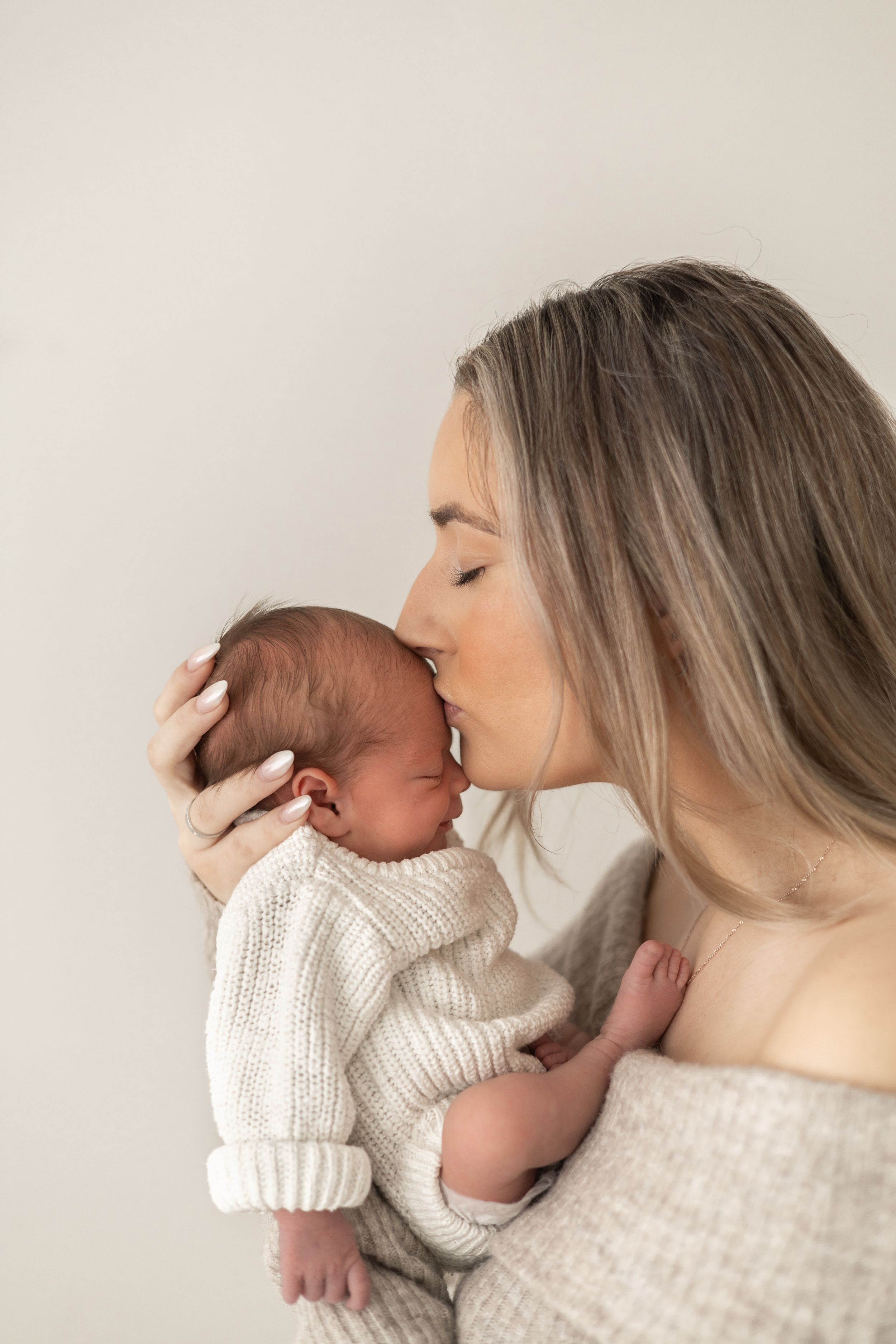 A young mother gently kisses her newborn baby against a neutral background newborn photographer Nottingham cosy home studio