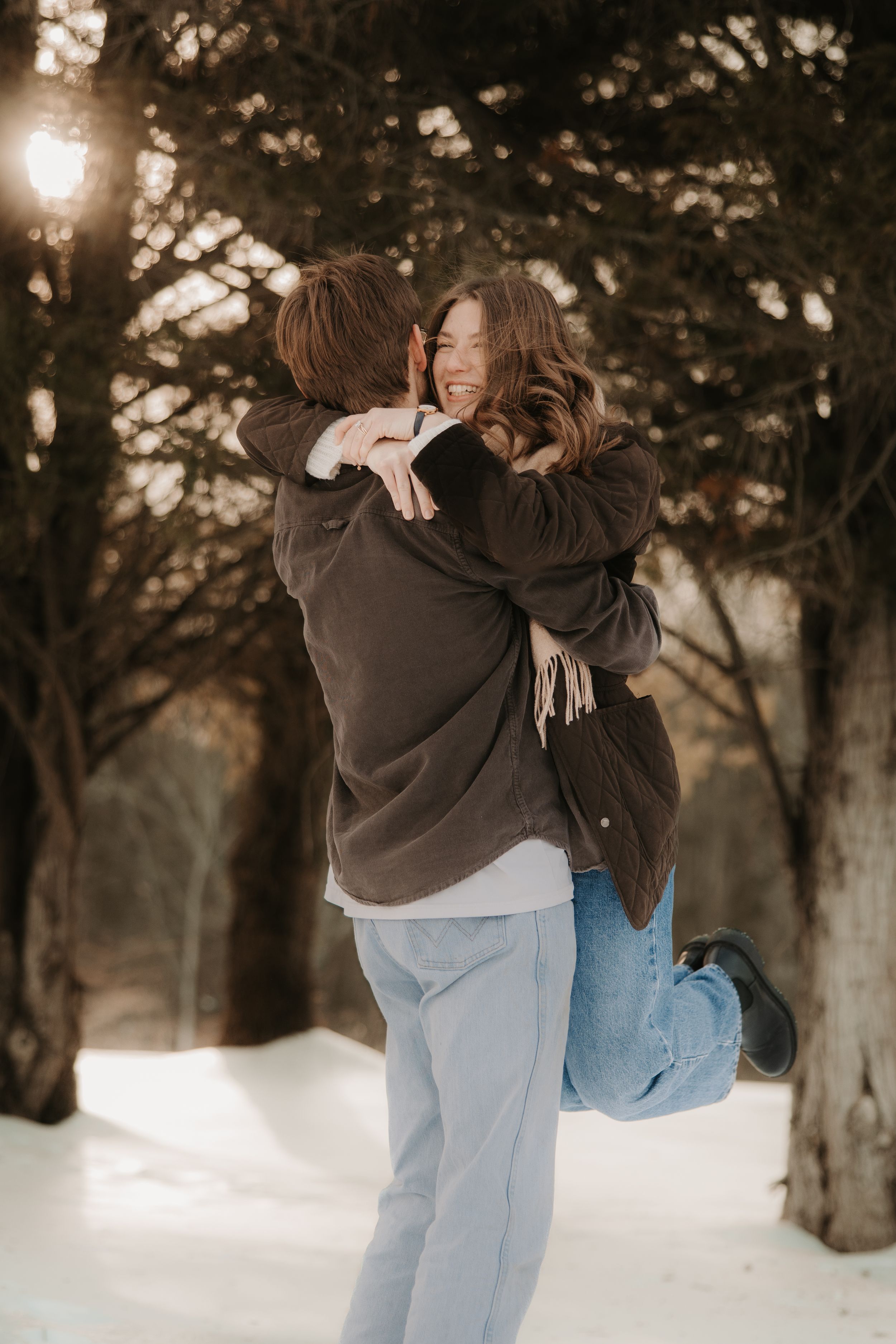 A couple embraces playfully in a snowy winter forest setting during golden hour.