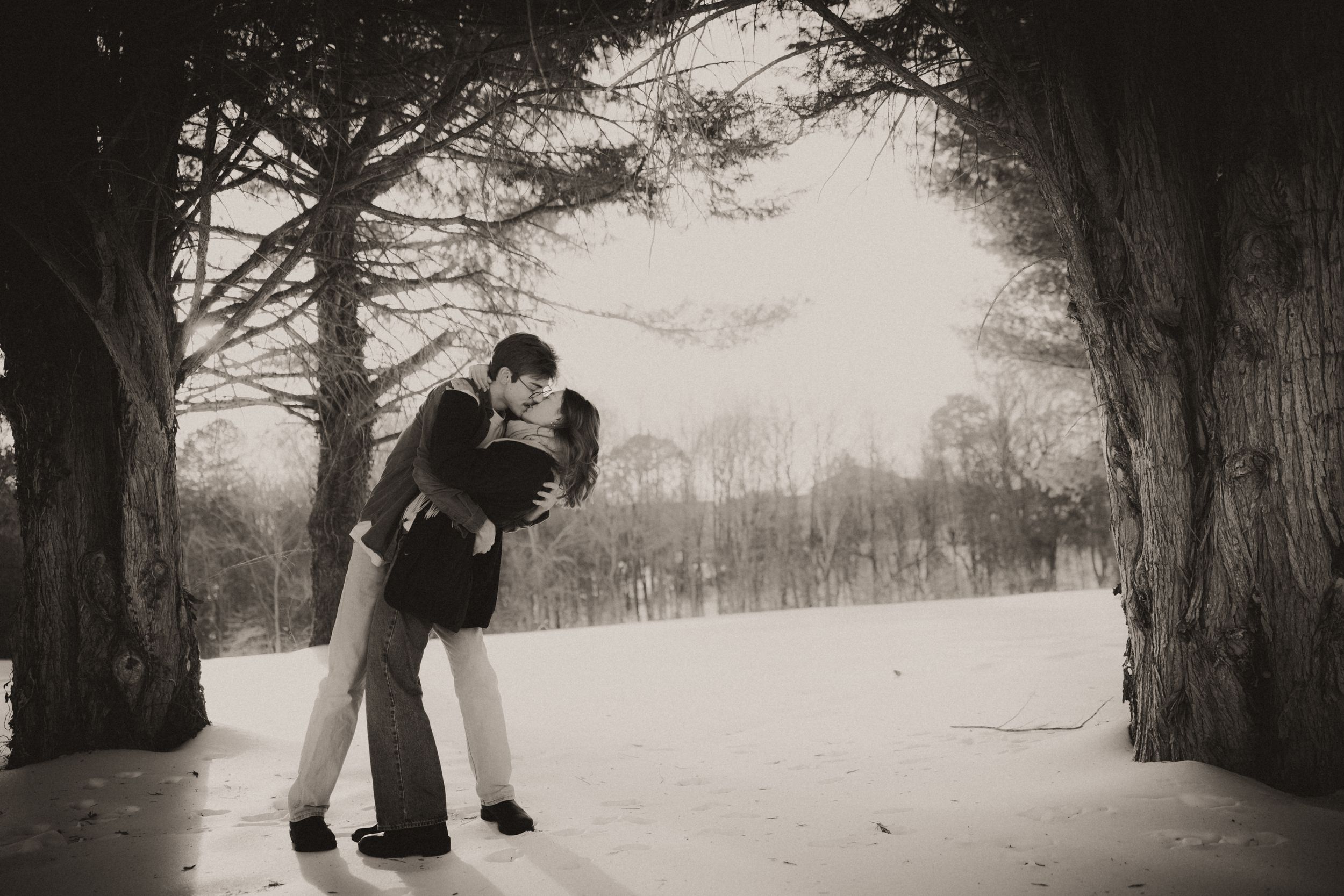A romantic couple embraces in a snowy winter landscape beneath tall evergreen trees.