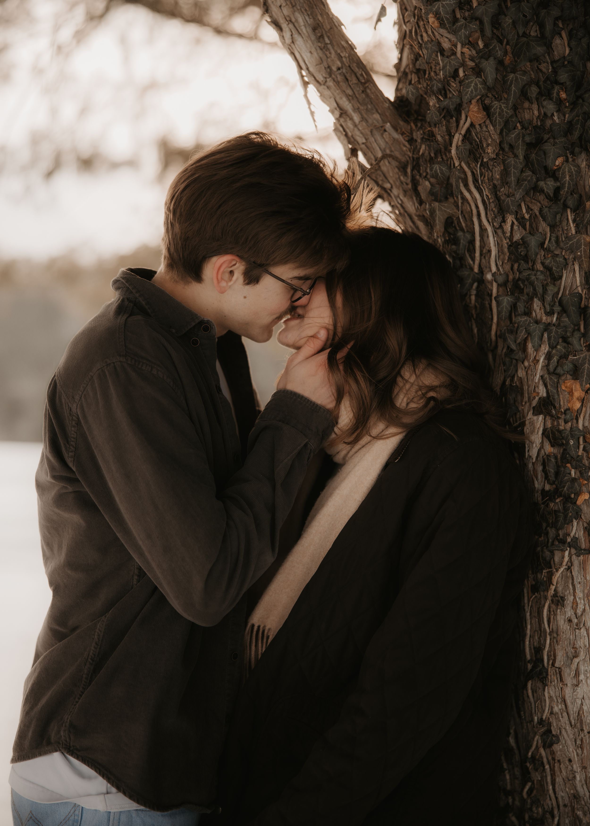 A romantic couple shares tender kisses against a tree trunk in a moody sepia-toned photo series.